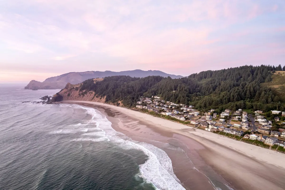A panoramic scene featuring rolling ocean waves, a sandy beach, a quaint town, and distant mountains. The beach appears inviting for various activities such as hiking and beachcombing, while the town conveys the allure of coastal communities like Depoe Bay and Newport.