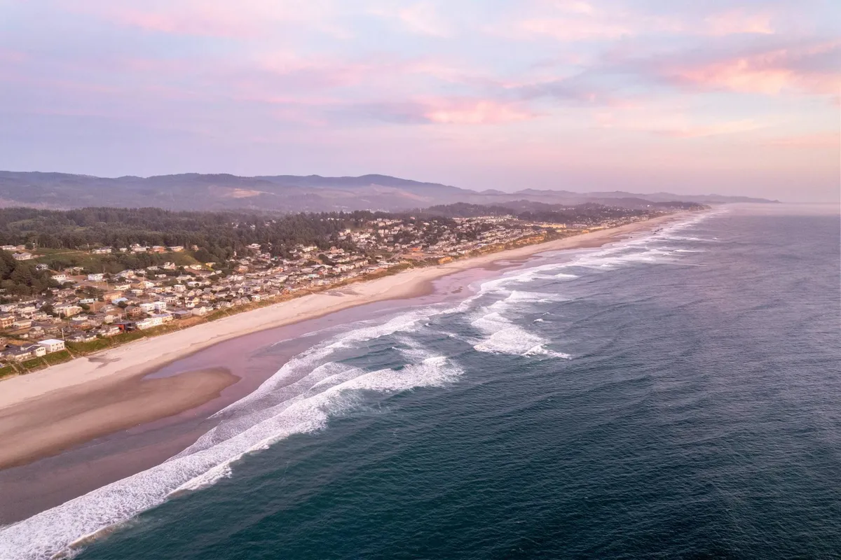 A broad view featuring ocean waves, the coastal town, mountain backdrops, and a colorful sunset sky. The composition captures the dynamic spirit of Lincoln City, evoking feelings of discovery and coastal adventure.