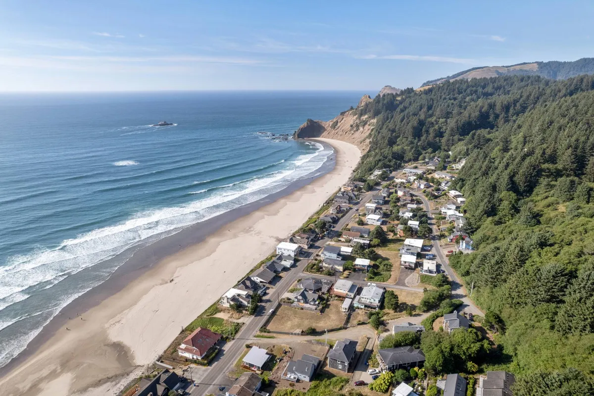 A stunning wide view of an expansive sandy beach bordered by waves, coastal cliffs, and a scattering of neighborhood houses. The natural shoreline stretches into the distance, inviting exploration and beachcombing, while the rocky outcroppings hint at tide pools that are perfect for marine life adventures.