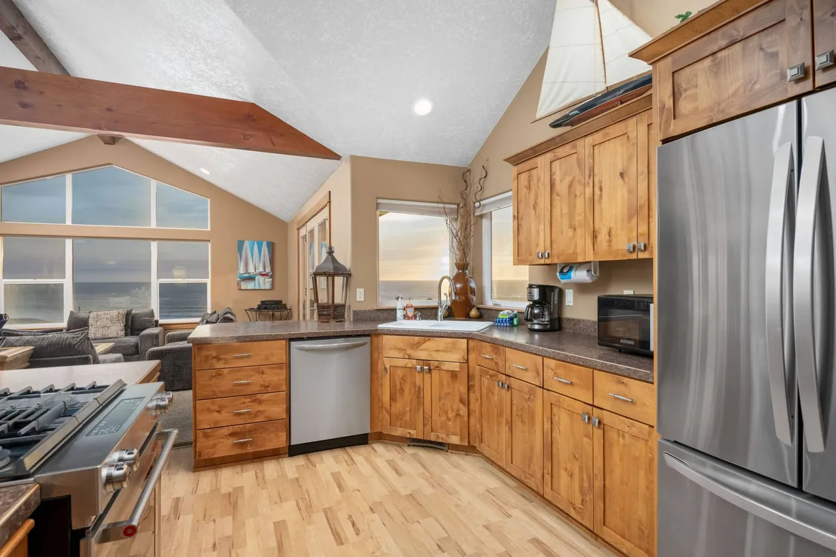 A stylish kitchen with stainless steel appliances, wooden cabinetry, and large windows filling the space with natural light. The open layout connects the kitchen to the living and dining areas, with decorative touches like a sailboat nodding to the coastal setting.