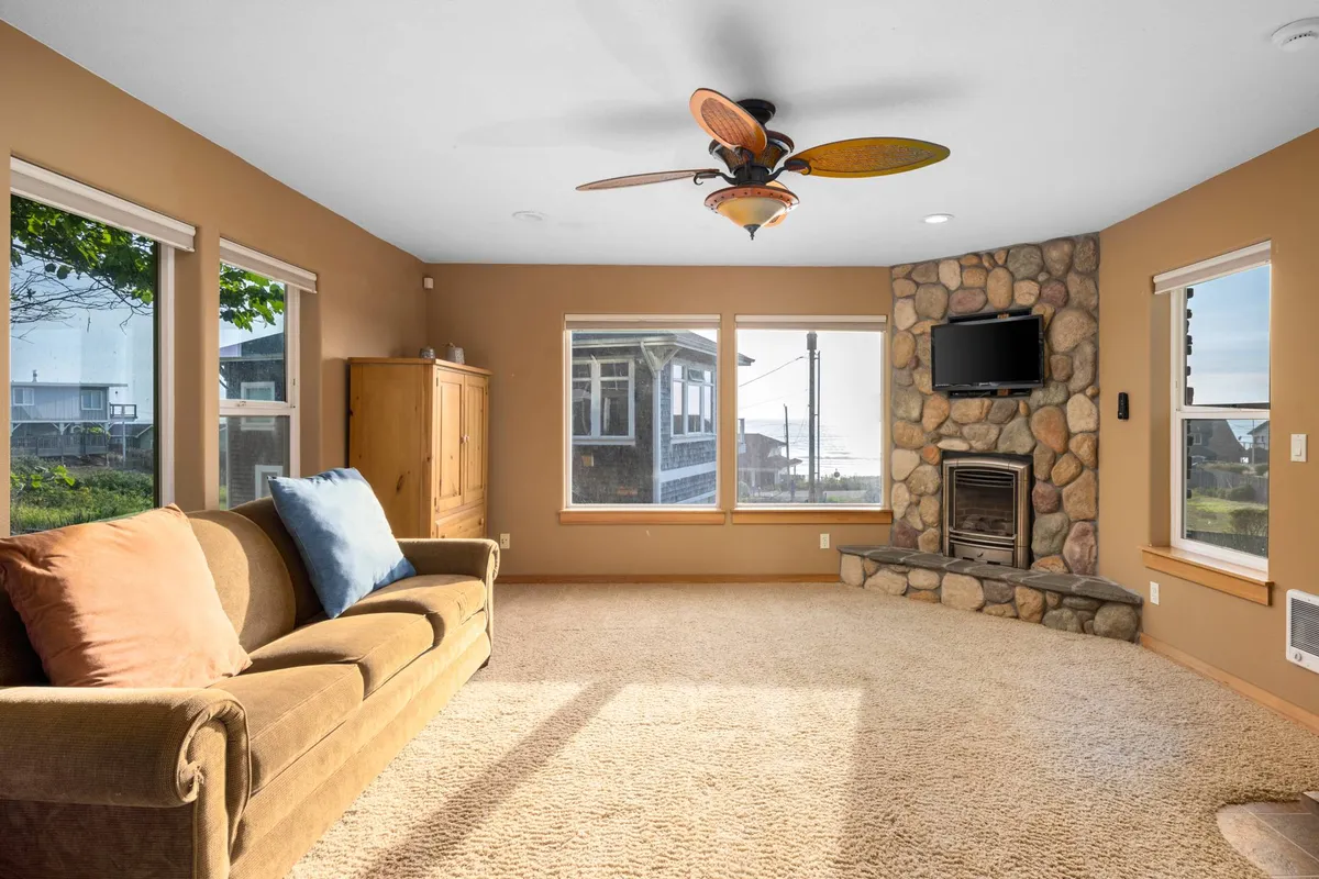 A cozy living room with a stone fireplace (clearly decorative), a plush sofa, large windows, and a television. Natural light fills the space, and the arrangement is ideal for relaxing, enjoying the view, or gathering for movie nights.