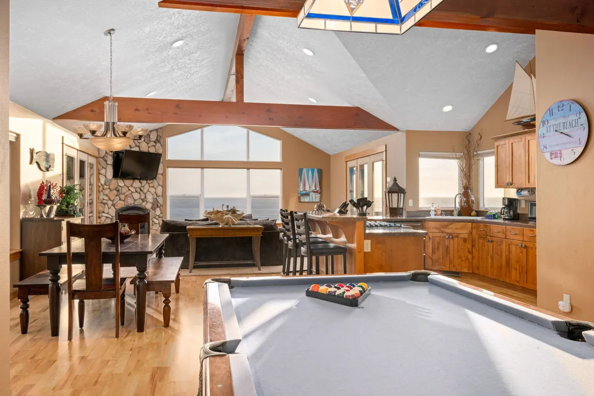 This image captures the heart of the gathering space: a bright, open-concept area featuring a pool table for game nights, a large wooden dining table for shared meals, and expansive windows framing sweeping ocean views. The kitchen area is visible, reinforcing the seamless flow between living, dining, and cooking spaces.