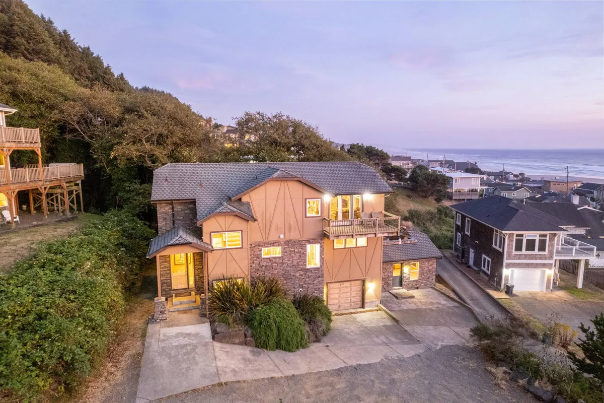 The image showcases the striking architectural design of a spacious, modern house surrounded by lush greenery, with sweeping ocean views visible in the background. The inviting balcony suggests a perfect spot for relaxing, while the peaceful setting highlights the home's tranquil atmosphere.