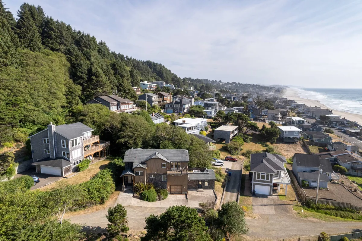 A sweeping view encompassing houses nestled between forested areas, a winding road, and the expansive ocean in the distance. The coastal landscape blends natural and community elements, with the shoreline visible, suggesting proximity to both natural attractions and local towns.