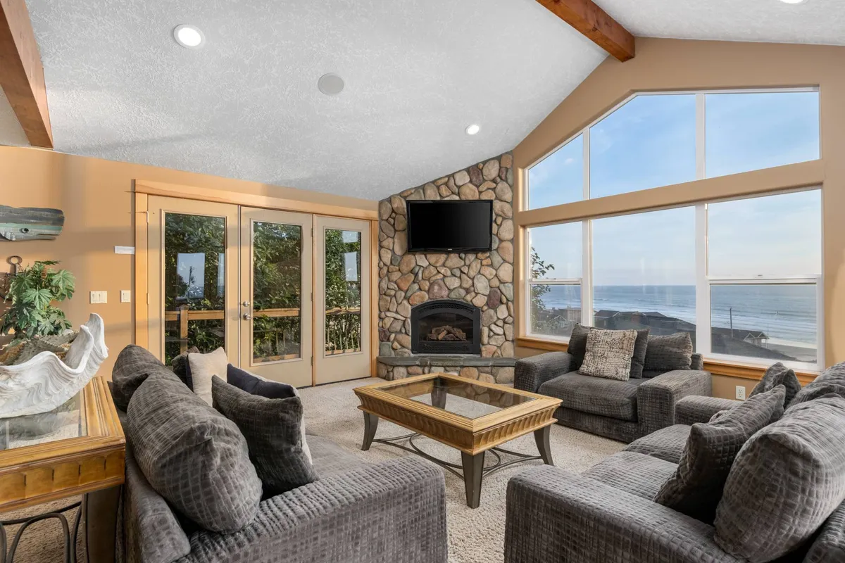 A cozy living room setup with comfortable seating arranged around a stone fireplace, set before large windows that showcase a stunning view of the Pacific. A coffee table and inviting atmosphere suggest relaxed gatherings and quiet moments watching the ocean.