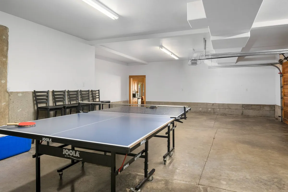 The garage game area is shown with a full-size Ping Pong table, stacked chairs, and the garage door visible, suggesting a fun, casual space for friendly matches and social gatherings.