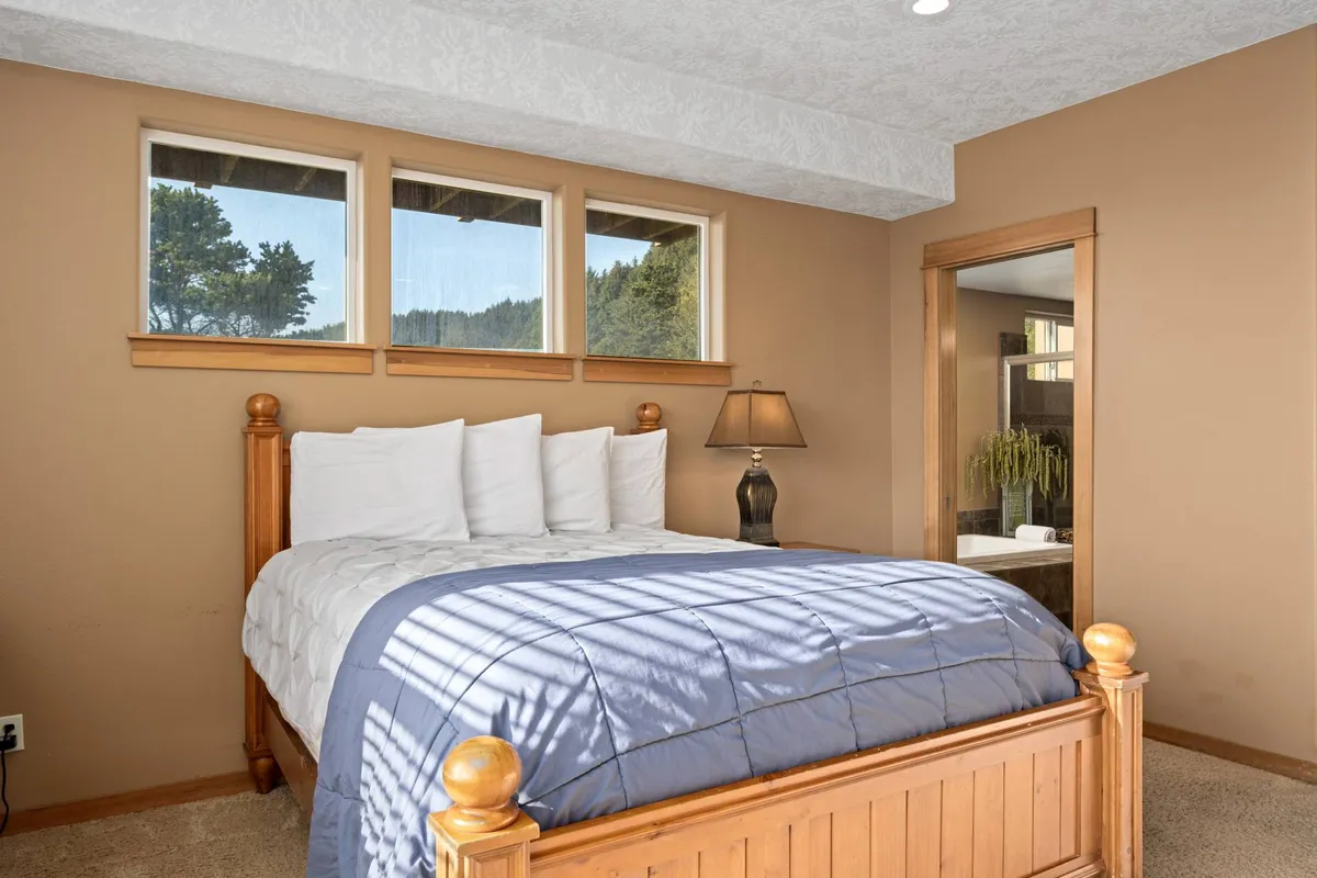 A serene bedroom with a plush bed, bedside lamp, and multiple windows letting in natural light. The simple, stylish decor promotes a sense of calm and relaxation, ideal for guests seeking comfort.